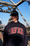 Person wearing a navy blue mockneck sweatshirt with 'SAVED' applique on it, standing in front of a bridge.