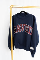 Navy blue sweatshirt with 'SAVED' text on a gold rack against a white background